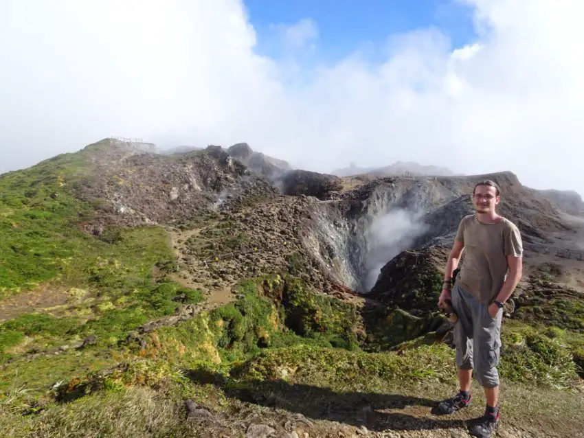 A man standing in front of a volcano crater spewing smoke