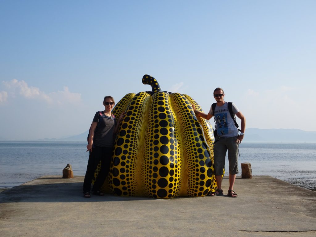 Two people standing next to a giant yellow pumpkin statue by the ocean