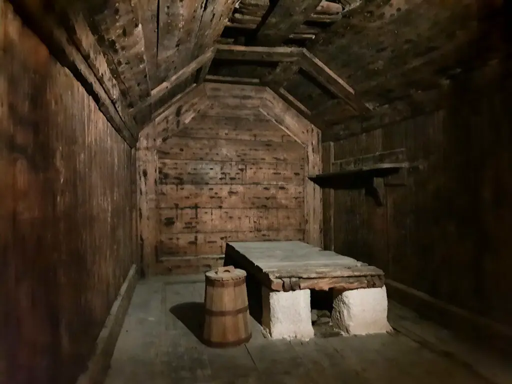 Asmall dark prison cell with a wooden cot and a bucket