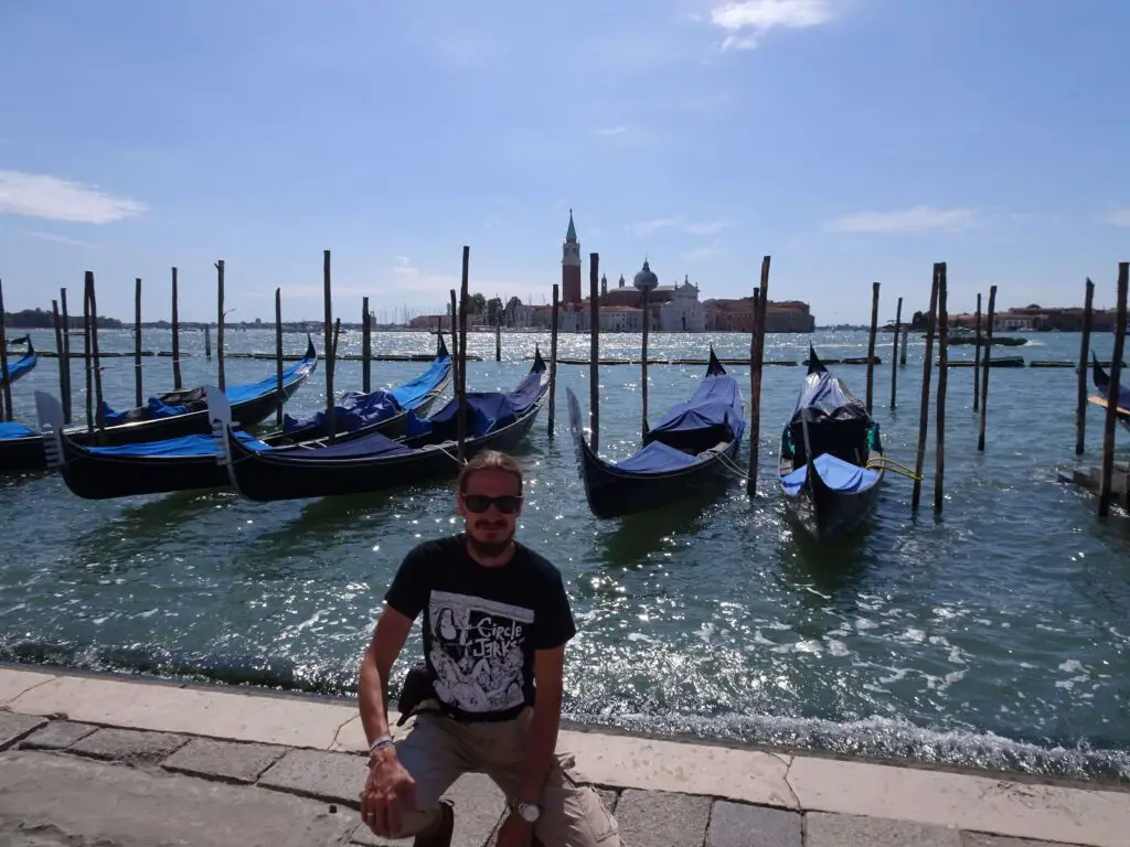 A man sitting at the water's edge in front of a row of gondolas with an island in the background