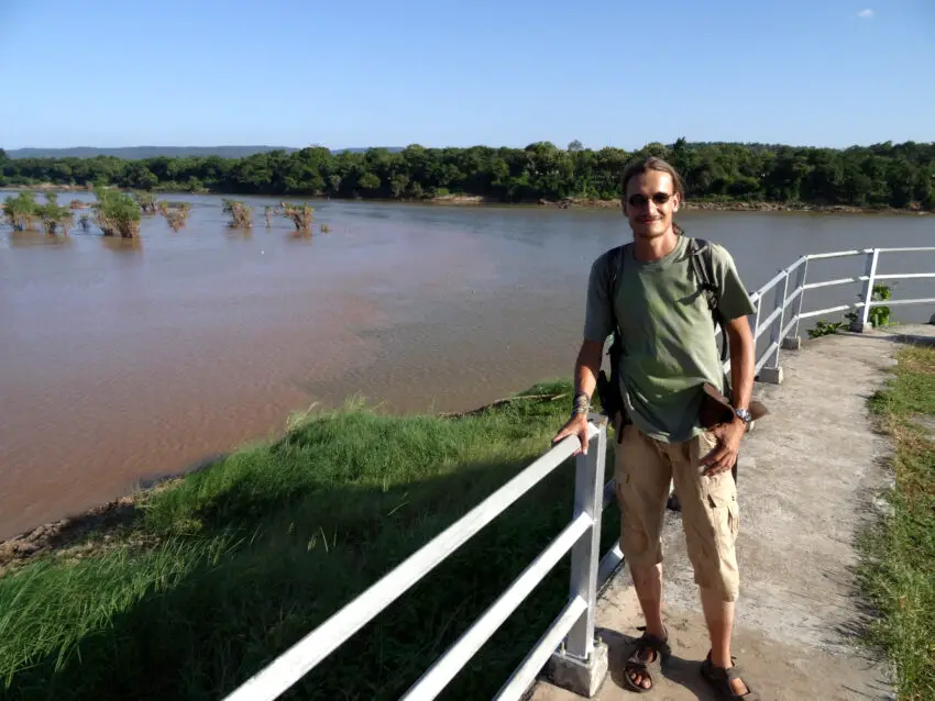 A man standing next to a river with a line of two distinctly coloured sections of water meeting down the middle