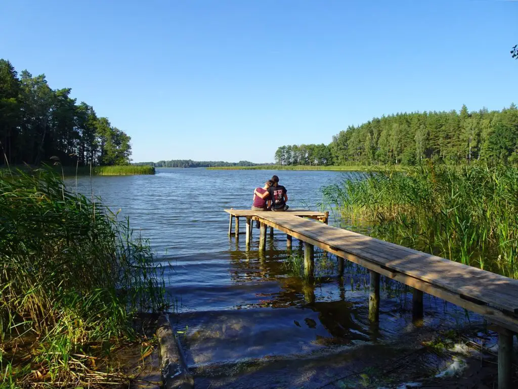 A man and woman sitting on a wooden pier over a lake, seen from the shore, facing away from the camera