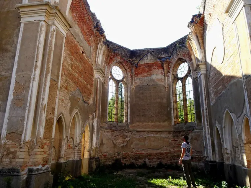 A man standing in the atmospheric ruins of a church built in a gothic revival style, looking at the walls of the apse