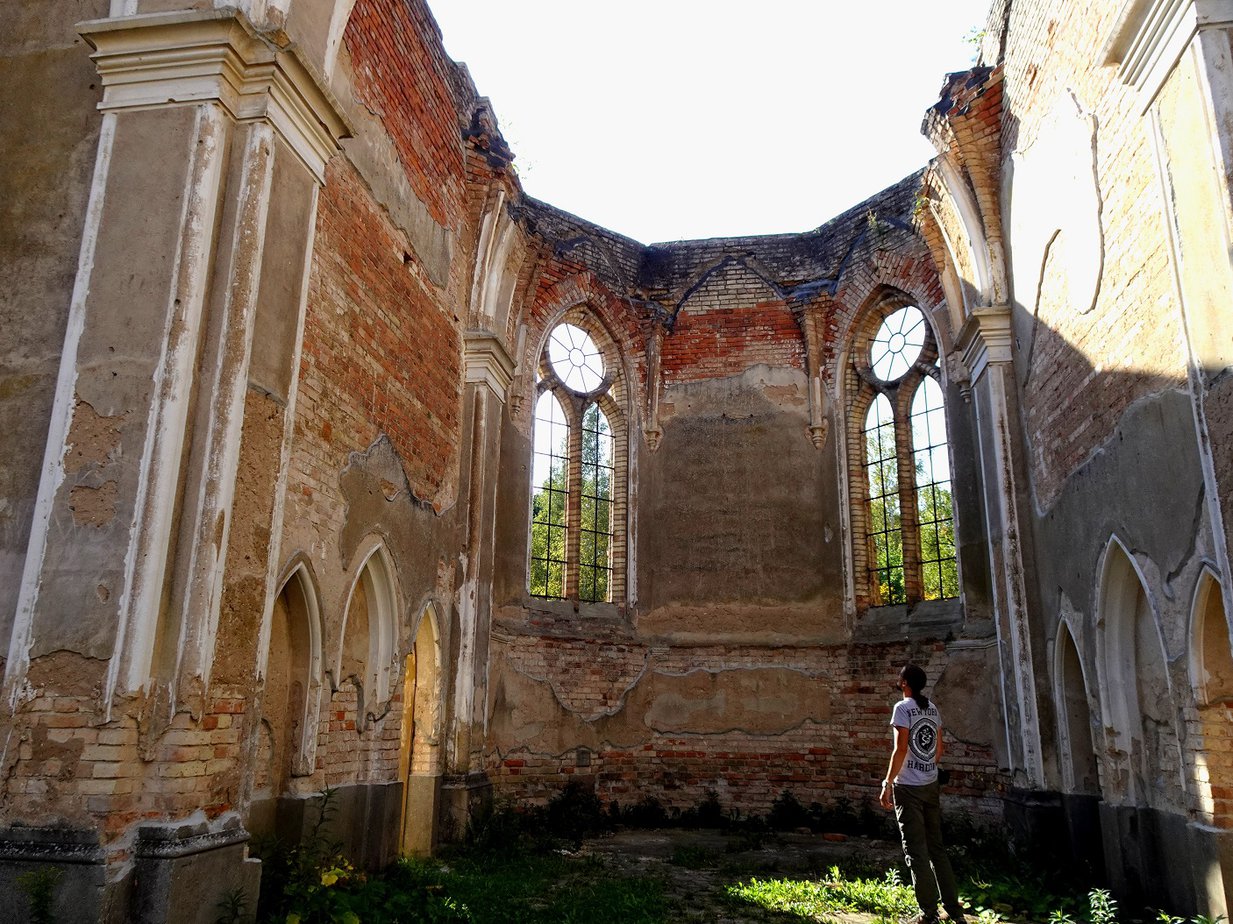A man standing in the atmospheric ruins of a church built in a gothic revival style, looking at the walls of the apse