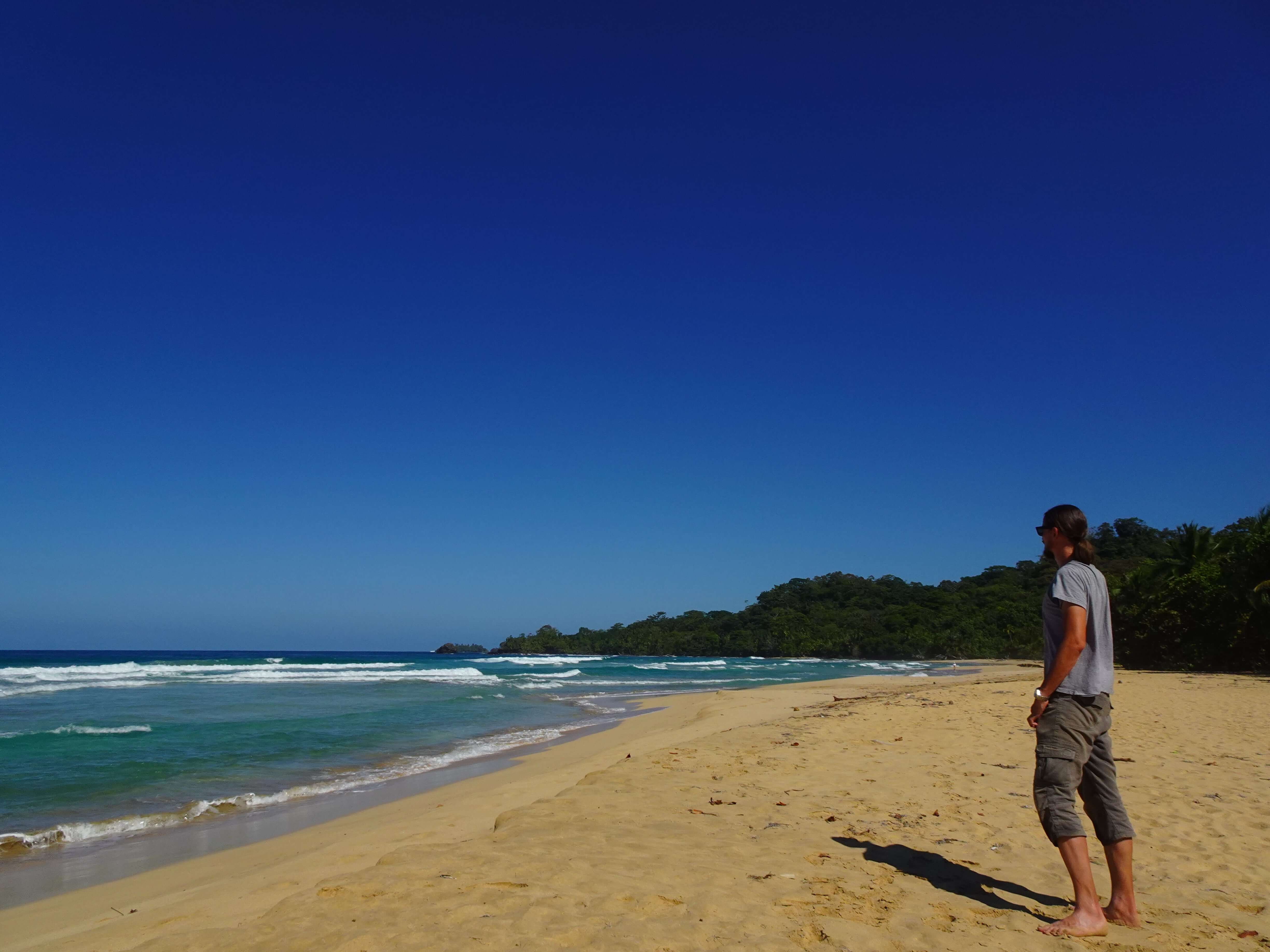 A man standing on a tropical beach, staring at the ocean.