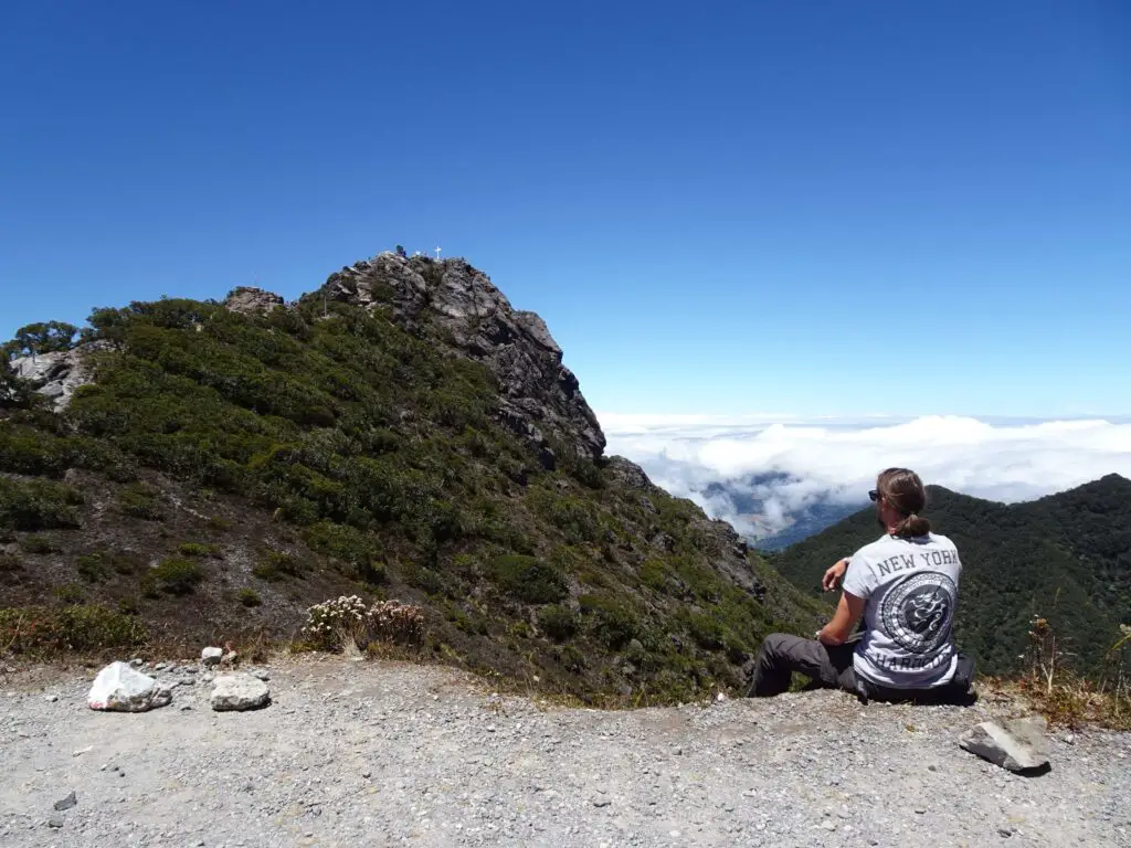 A man seen from behind, sitting on a mountain edge, looking at the rocky top of a mountain