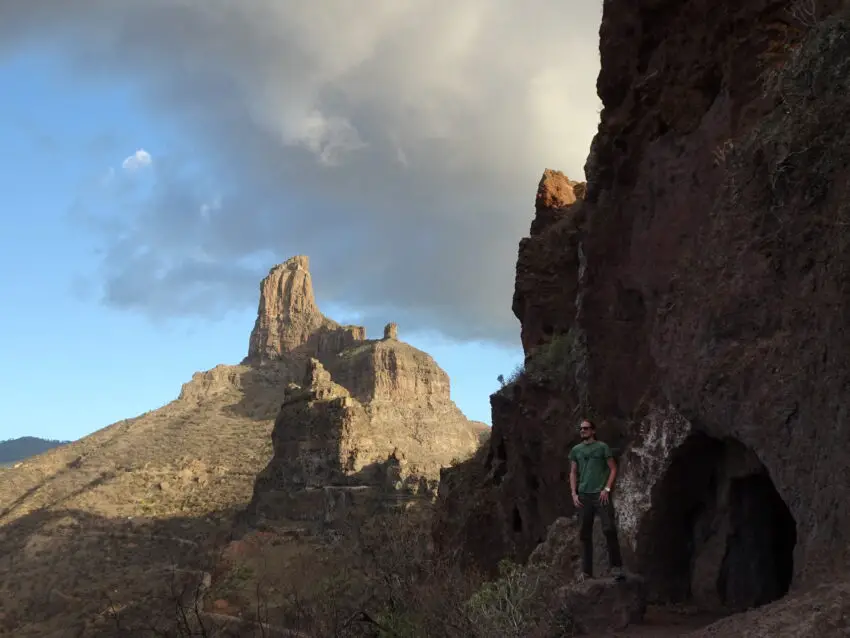 A man standing near a cave with a series of rock outcrops in the background