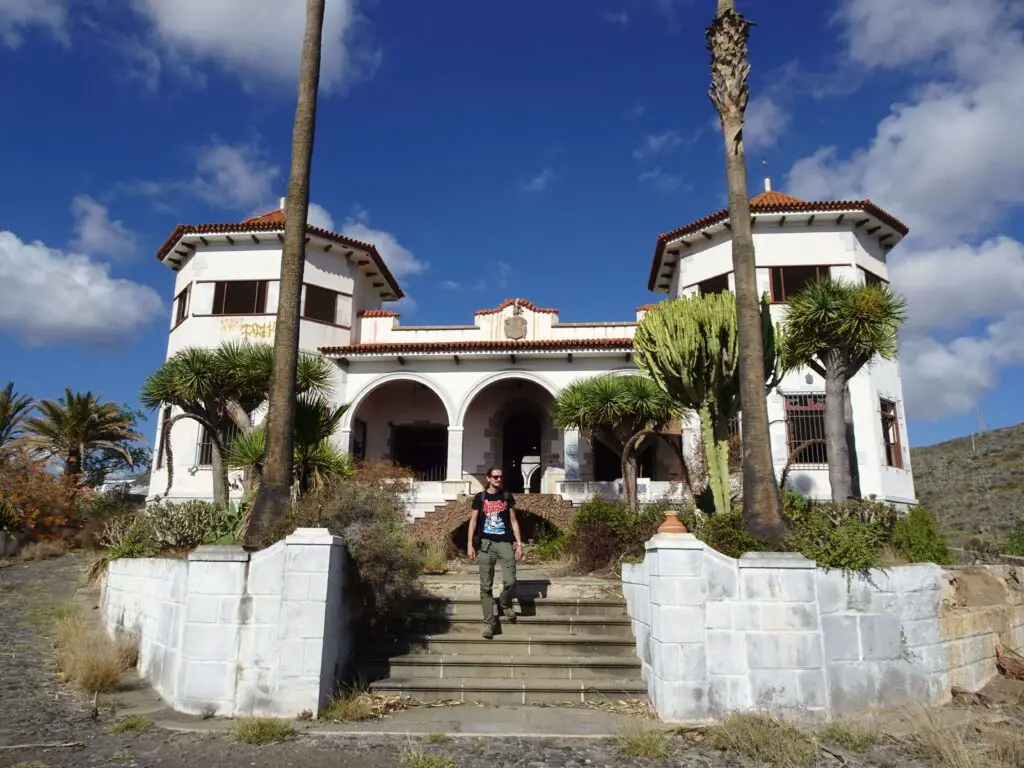 A man walking down the stairs in front of an abandoned villa framed by palm trees