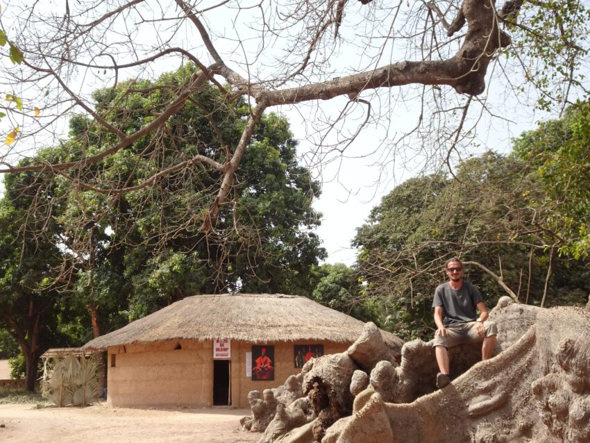 A man sitting on tall roots with an earthen roundhouse in the background