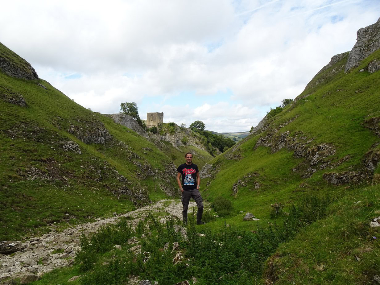 A man standing in a narrow valley with rocky slopes on both sides and a ruined stone tower atop one of them