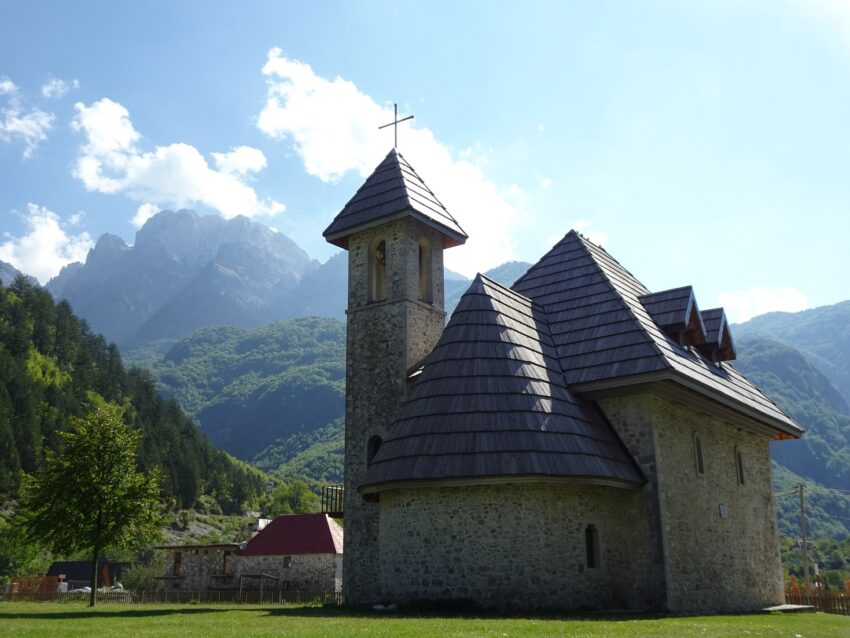A stone church ona sunny meadow with tall mountains in the background