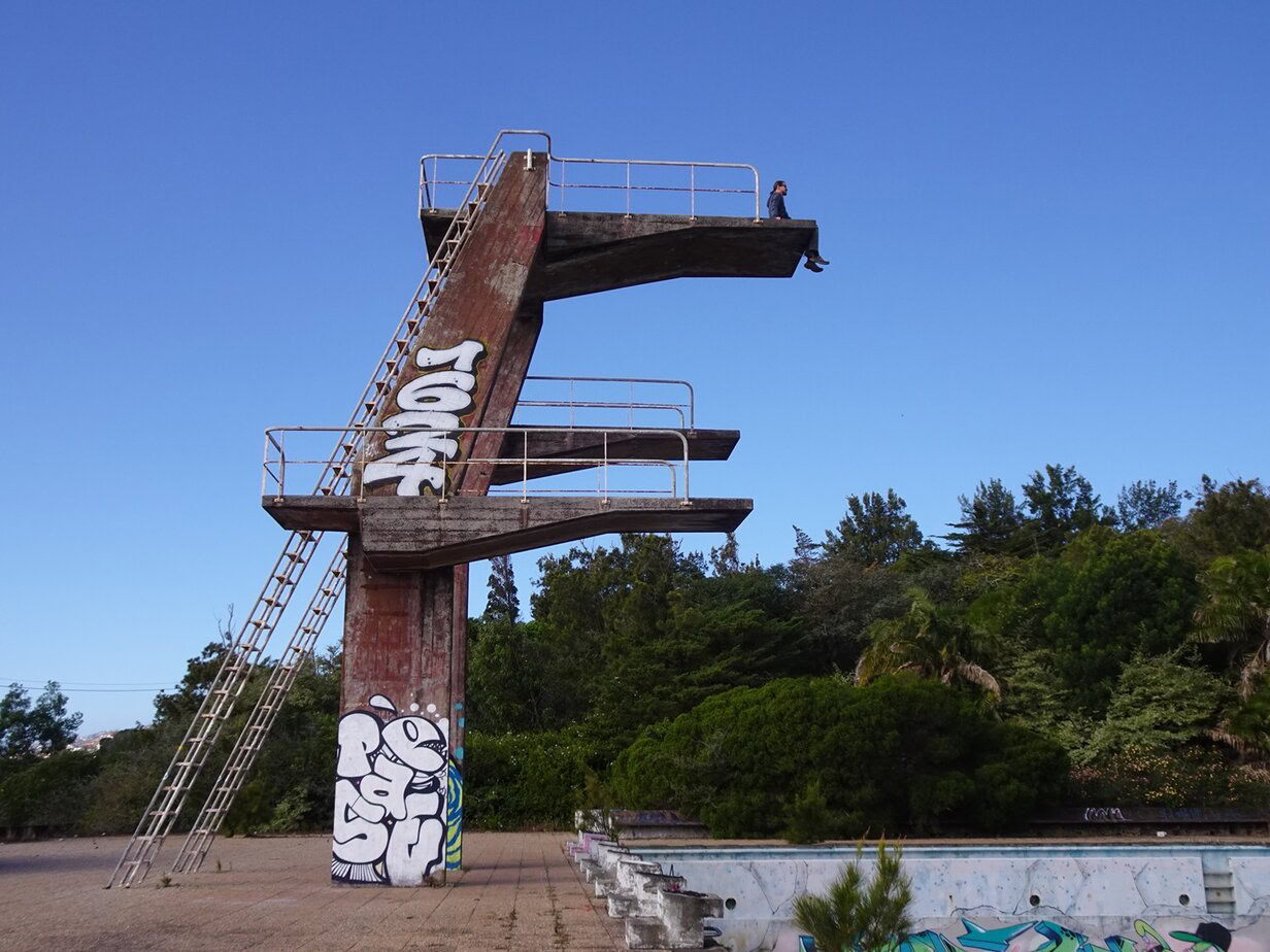 A man sitting at the edge of a diving tower over an empty swimming pool