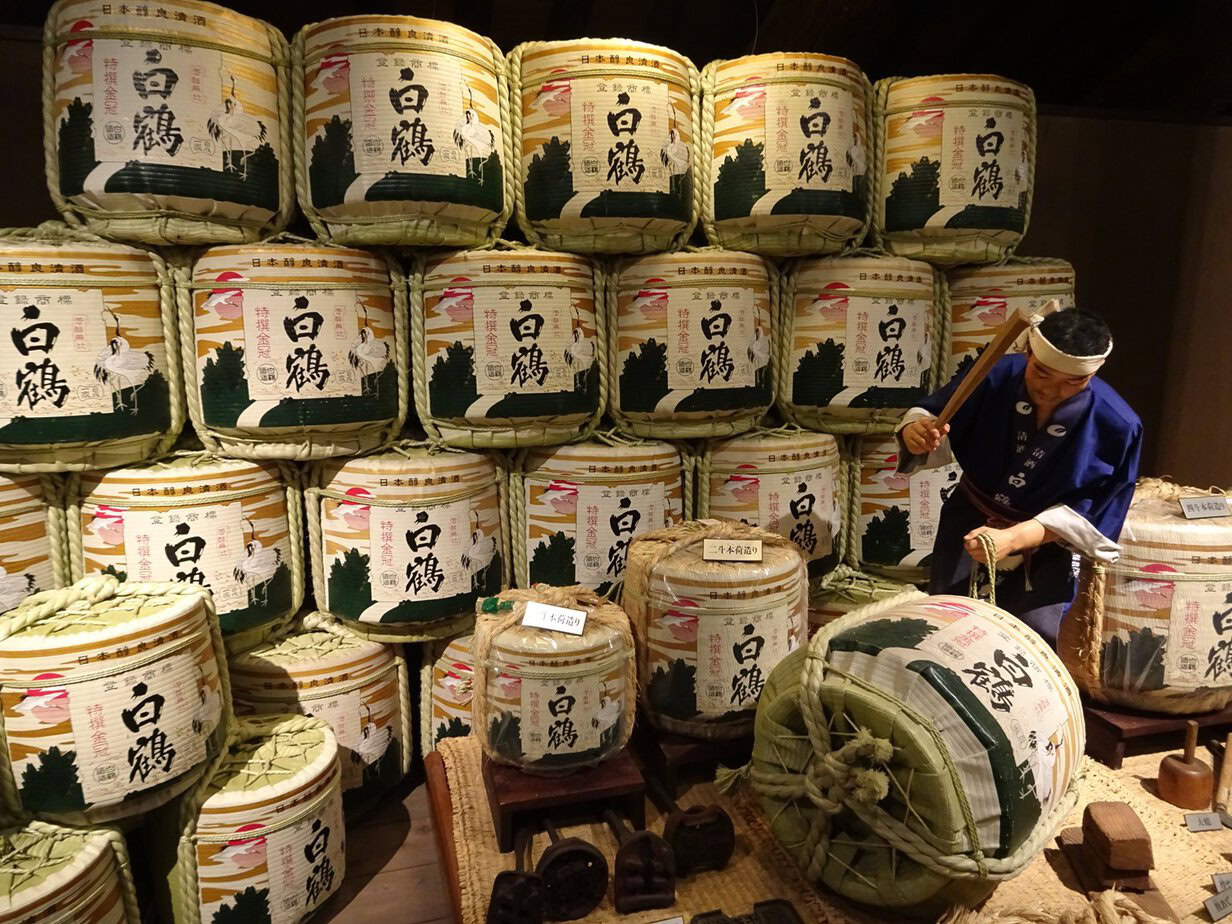 A museum display of sake barrels with a wax figure hammering on one
